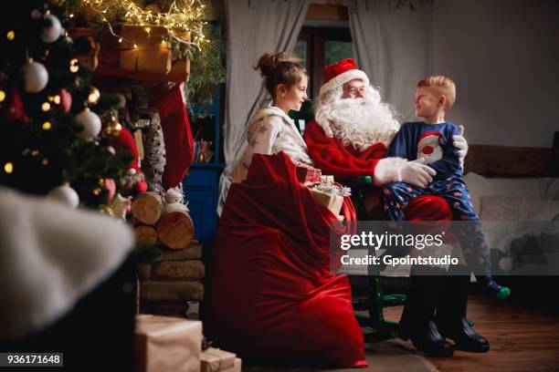 young girl and boy sitting with santa, sack full of presents beside him - grotte stock-fotos und bilder