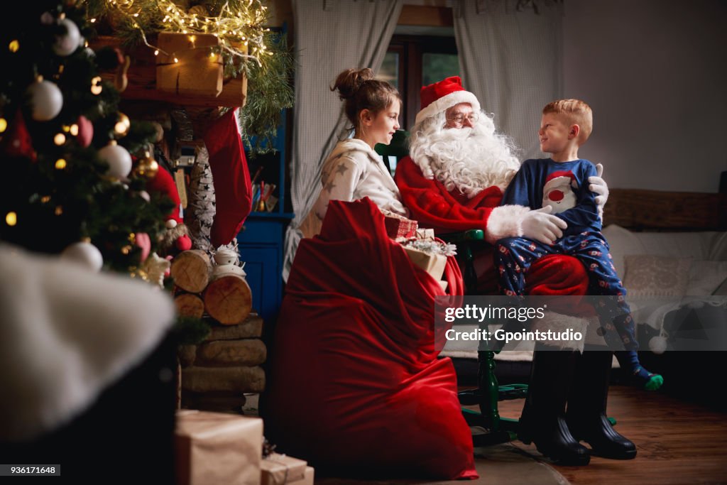 Young girl and boy sitting with Santa, sack full of presents beside him