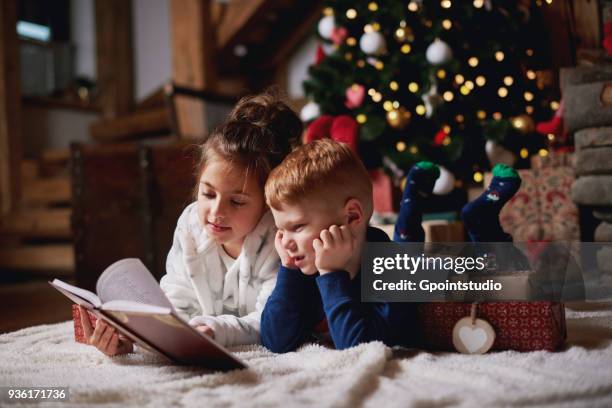 young girl and boy relaxing beside christmas tree, reading book - children only stock pictures, royalty-free photos & images