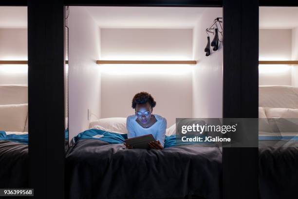 young adult woman lying down using a digital tablet in a capsule hotel - capsule hotel stock pictures, royalty-free photos & images