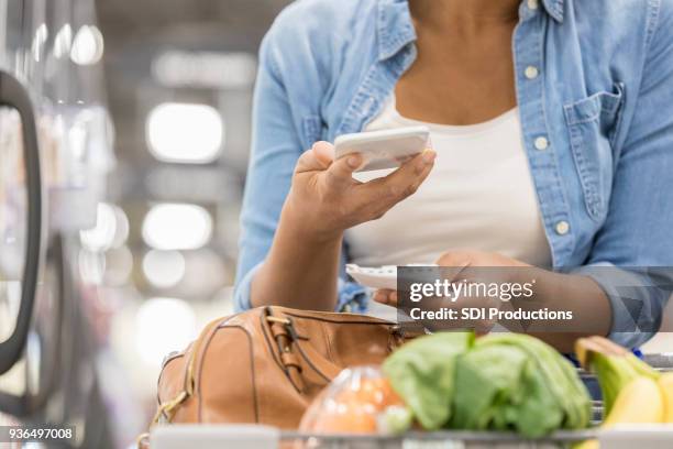 unrecognizable supermarket customer uses smart phone and paper list - woman shopping list stock pictures, royalty-free photos & images