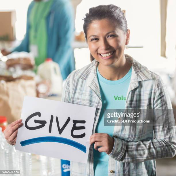 cheerful woman holds "give" sign in local soup kitchen - brand advocacy stock pictures, royalty-free photos & images