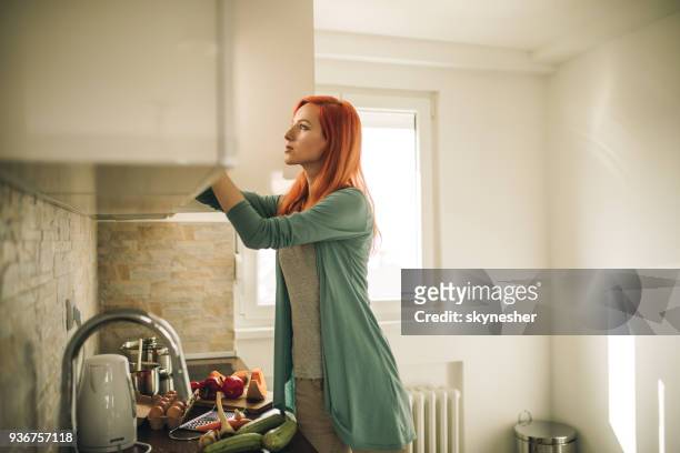 redhead woman searching for something in kitchen cabinet. - beauty cabinet stock pictures, royalty-free photos & images