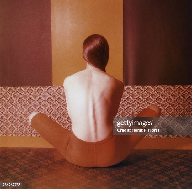 Back of a topless model sitting with her knees spread on a patterned tile floor wearing a red Jantzen knee-length panty control garment.