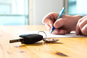 Man signing car insurance document or lease paper. Writing signature on contract or agreement. Buying or selling new or used vehicle. Car keys on table. Warranty or guarantee.