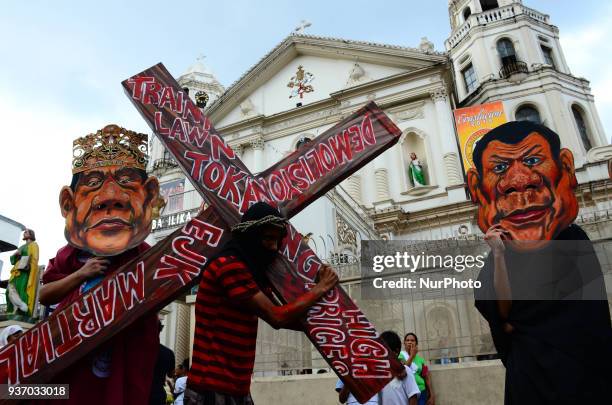 An activist carrying a cross stands in between other activists wearing Duterte masks. Various groups gather at Plaza Mirada on March 23, 2018 for a...