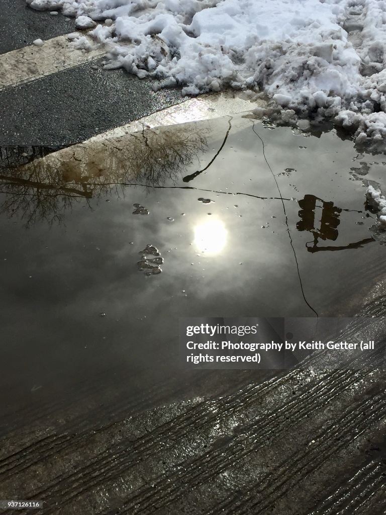 View of a street corner after a snowstorm with snow, slush & melting water reflecting the sky and street signals above.