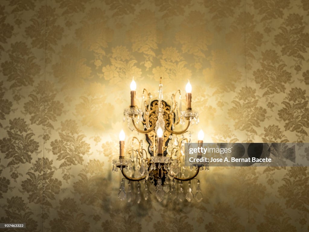 Close-up of crystal wall lamp of classic style with the flushed lights on a wall with 	wallpaper.