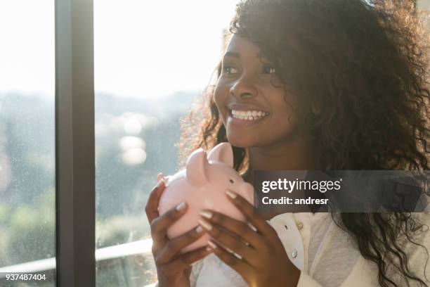 schöne schwarze lateinamerikanische frau hält ihr sparschwein tagträumen neben dem fenster - african american woman with money stock-fotos und bilder