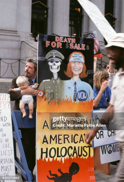 New York, New York, USA - July 13 to July 16, 1992: A man holds a baby doll next to a large anti-abortion poster saying 'Death for $ Sale $" above a...