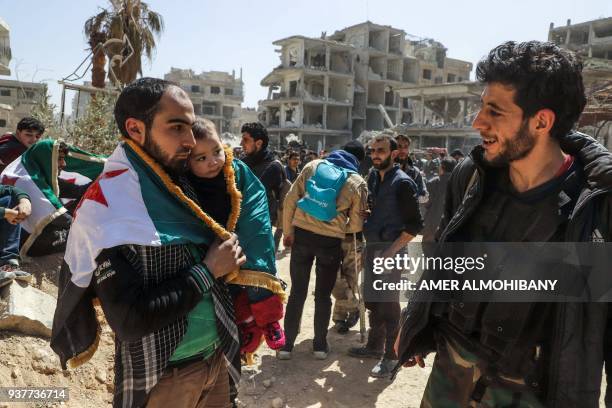 Syrian man carrying a child and draped in the flag of the opposition talks to another as they wait for evacuation from the town of Arbin in the...