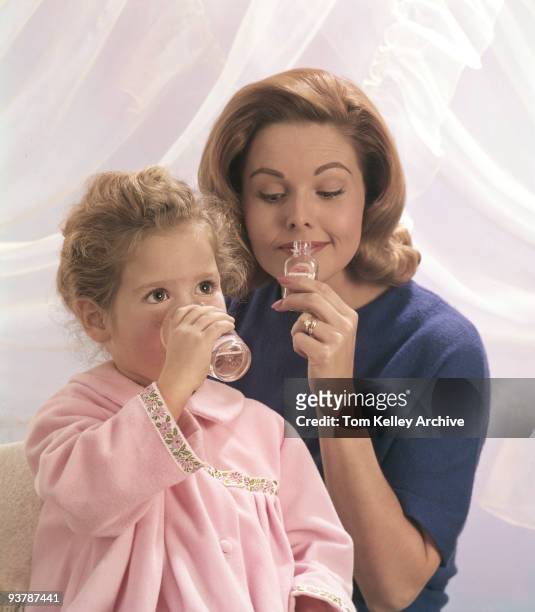 Little gril drinks from a clear glass while a women holds a small bottle of medicine up to her own mouth, 1964.