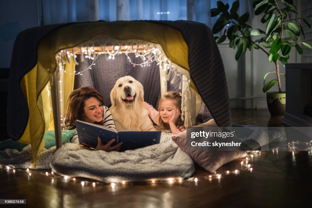 Happy mother and daughter relaxing with a dog in a tent and reading a book.