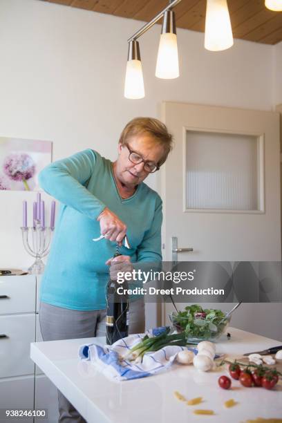 old woman opening a bottle of red wine in the kitchen - brille frau stockfoto's en -beelden