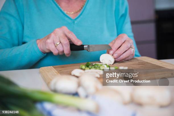 old woman cutting mushrooms and spring onions at the kitchen table - menschliches körperteil foto e immagini stock