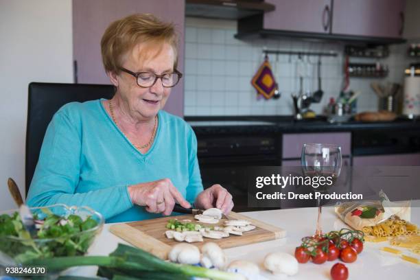 old woman cutting spring onions and button mushroom at the kitchen table - organisch fotografías e imágenes de stock