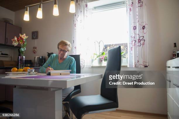 old woman watching digital tablet and writing in book at dining table - frühstück tisch stock pictures, royalty-free photos & images