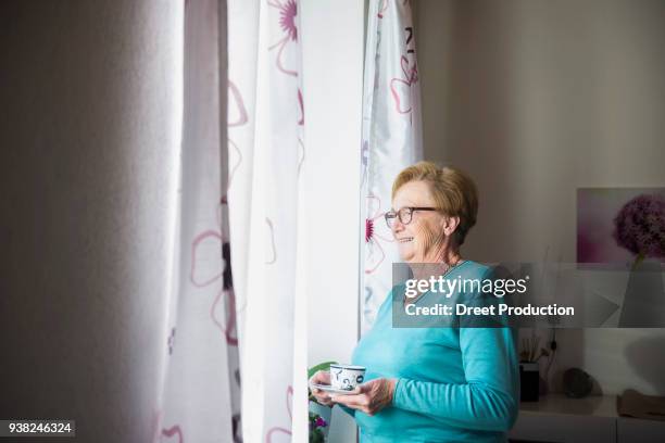 smiling old woman looking out of window with a cup of coffee - eine frau allein fotografías e imágenes de stock