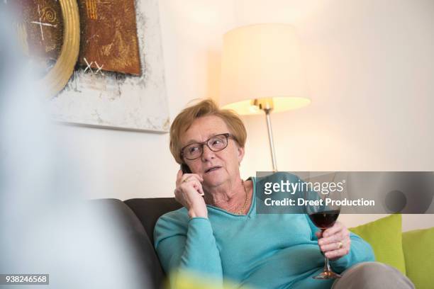 old woman relaxing on sofa with a glass of red wine and talking on phone - wohnzimmer photos et images de collection