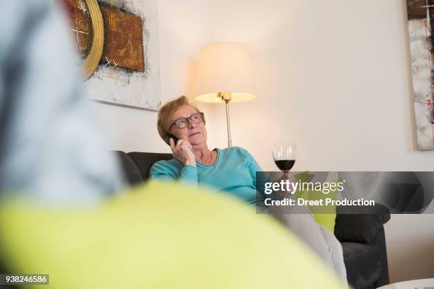 old woman relaxing on sofa with a glass of red wine and talking on phone - weinglas stockfoto's en -beelden