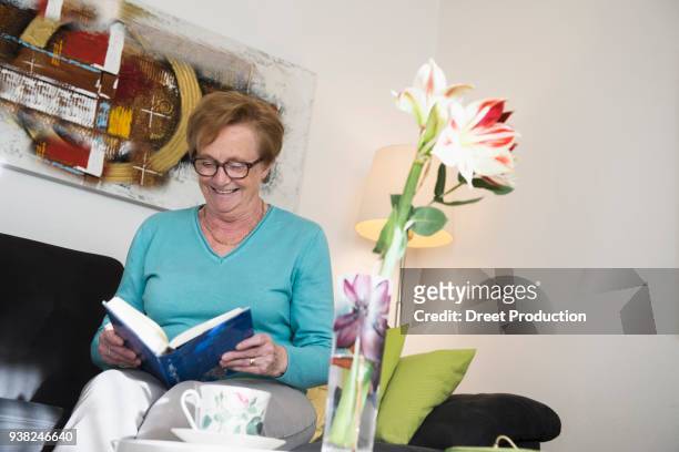 happy old woman sitting on sofa reading a book - wohnzimmer couch stock pictures, royalty-free photos & images