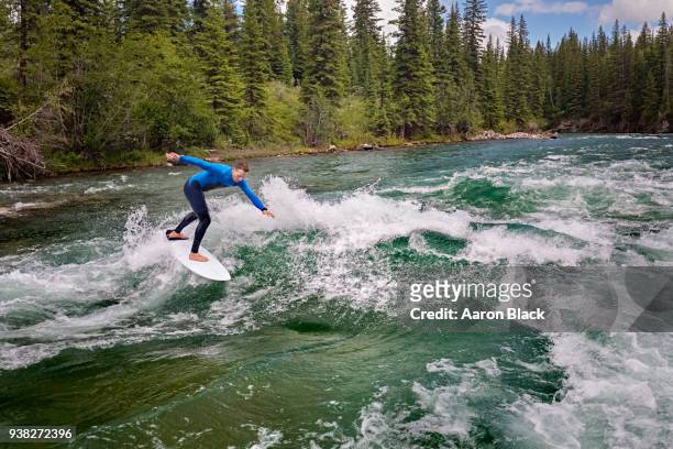 man in a blue wetsuit standing on surfboard surfing a turquoise wave in the middle of a river. - kananaskis stock pictures, royalty-free photos & images