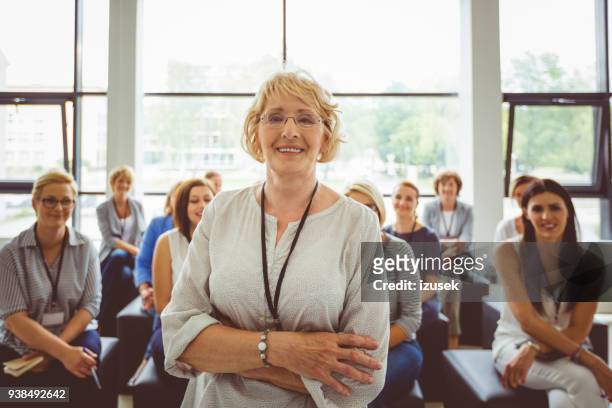 porträt von lächelnden senior weibliche moderatorin mit publikum im hintergrund - education retirement stock-fotos und bilder