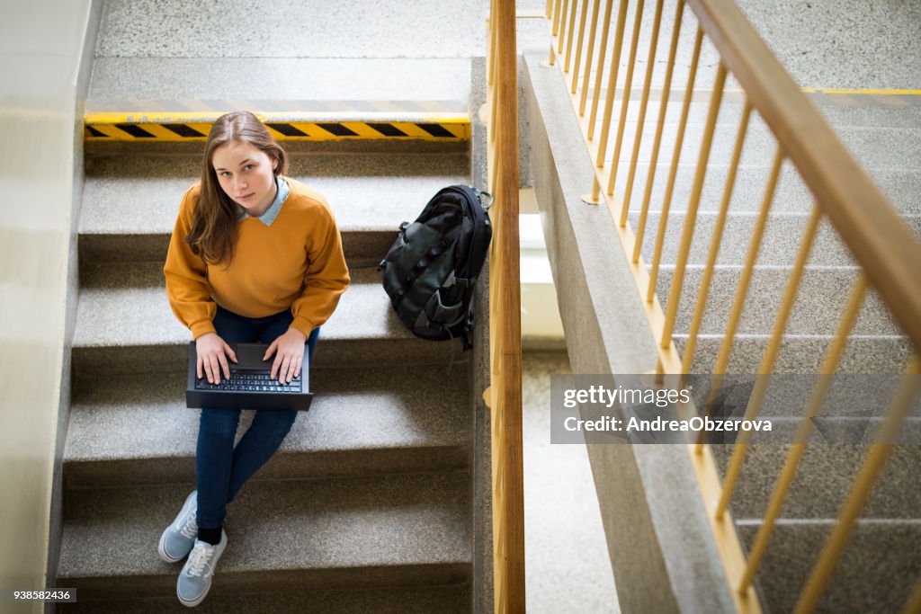Young female college student sitting on stairs at school, writing essay on her laptop and looking up at the camera. Education concept. View from above.