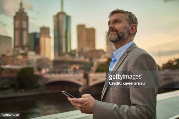 businessman holding phone at dusk in melbourne centre city - melbourne australia stock pictures, royalty-free photos & images