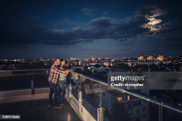 amorosa pareja que está parado en un balcón del penthouse de noche. - moonlight lovers fotografías e imágenes de stock