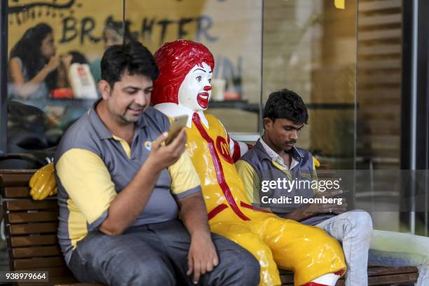 Food delivery couriers look at smartphones while sitting next to a statue of Ronald McDonald outside a McDonald's Corp. Restaurant, operated by...