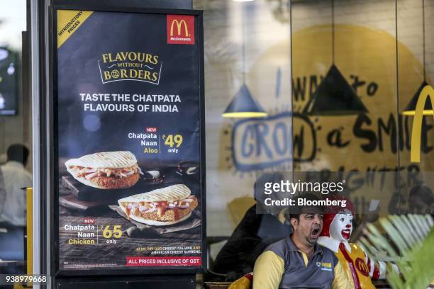 Food delivery courier yawns while sitting next to a statue of Ronald McDonald as menu items are displayed in the window of a McDonald's Corp....
