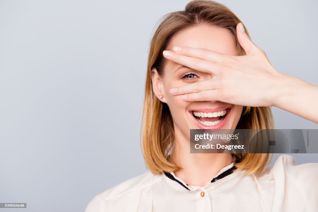 Blond young woman with beaming smile is looking through her hand. She is wearing casual outfit and stands on light grey background