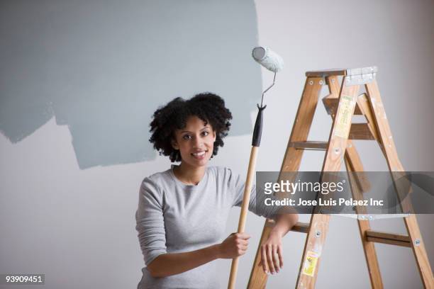 mixed race woman holding paint roller and leaning on ladder - wand streichen stock-fotos und bilder