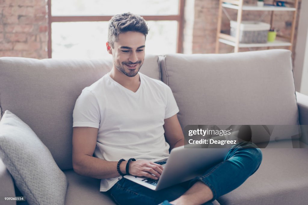 Young attractive smiling guy is browsing at his laptop, sitting at home on the cozy beige sofa at home, wearing casual outfit