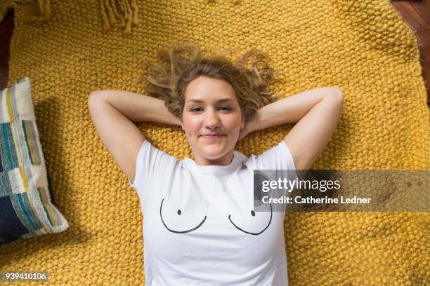 young blonde haired woman with a boob t-shirt lying on a yellow rug and smiling. - seno fotografías e imágenes de stock