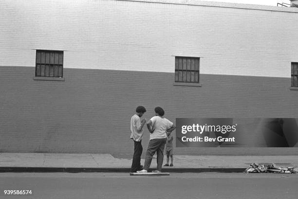 Two young men, accompanied by a shirtless boy, talk together on a sidewalk in the El Barrio neighborhood, East Harlem, New York, New York, July 1969....