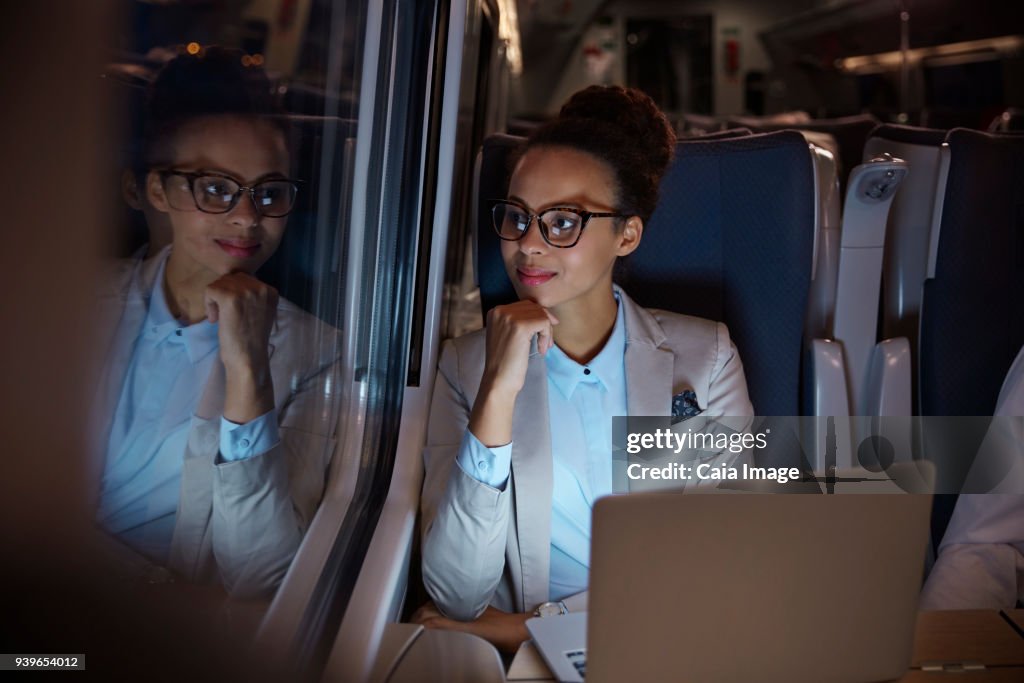 Confident, thoughtful businesswoman looking out window on passenger train at night, working at laptop
