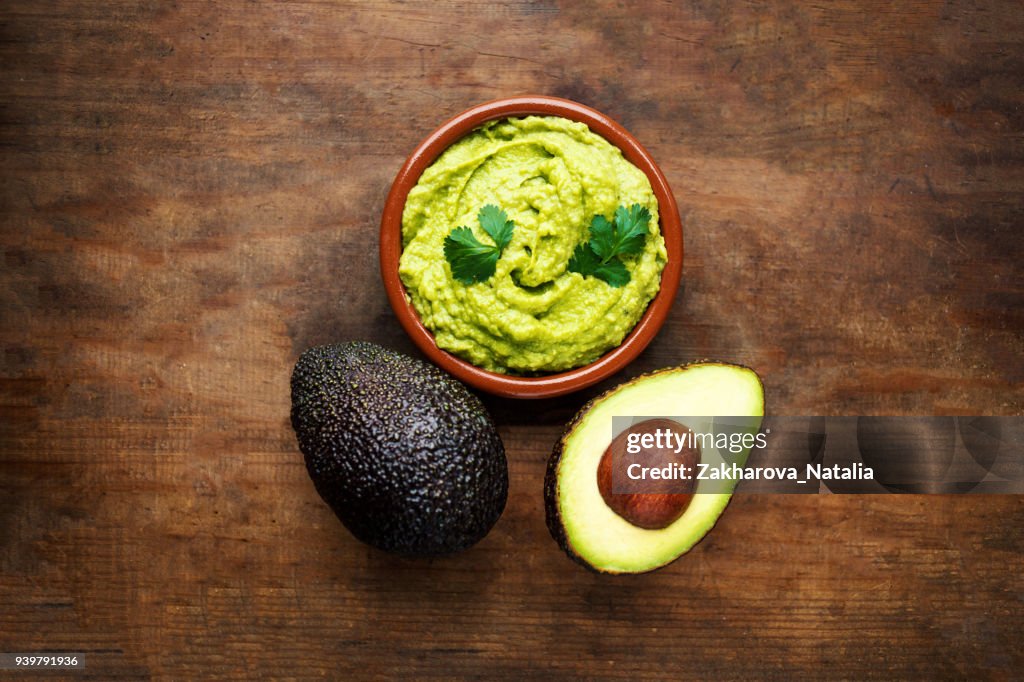 Avocado with guacamole sauce on a dark wood background. Half and whole avocadoes close up. Top view. Copy space"n
