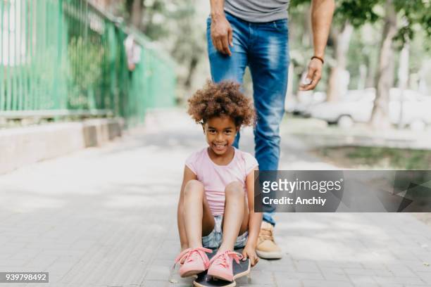 happy african ethnicity girl skateboarding in the park with her dad - father longboard stock pictures, royalty-free photos & images