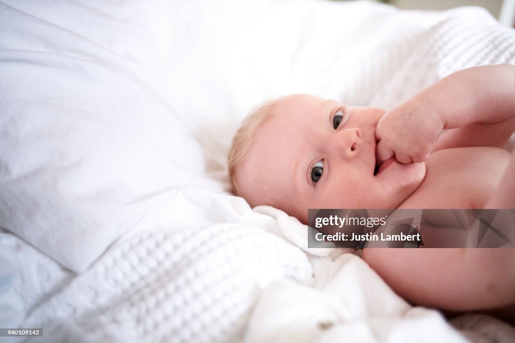 Baby girl lying on parents bed with fingers in mouth looking to camera