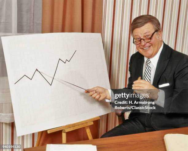 1970s HAPPY BUSINESSMAN LOOKING AT CAMERA POINTING TO GRAPH CHART INDICATING RISE IN BUSINESS SALES PROFITS