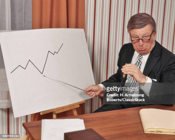 1970s UNHAPPY BUSINESS MAN LOOKING AT CAMERA POINTING TO GRAPH CHART INDICATING A DECLINE IN SALES PROFITS BUSINESS