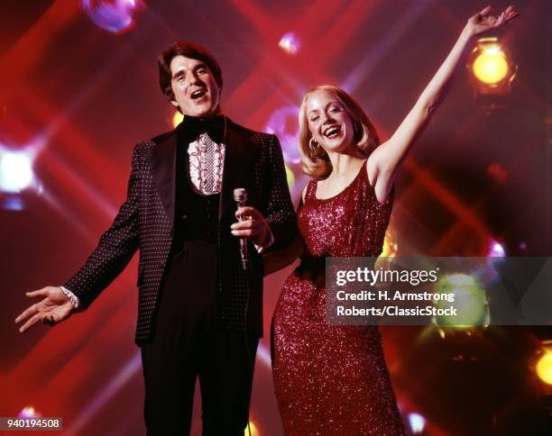 1970s WOMAN AND MAN WITH MICROPHONE SINGING AND GESTURING STROBE LIGHTS IN BACKGROUND TUXEDO GOWN ENTERTAINING