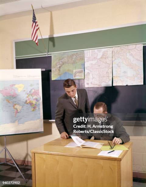 1960s 2 MEN TEACHER AT DESK TEEN STUDENT STANDING LOOK AT PAPERS WEAR SUIT & TIE CHALK BOARD MAPS FLAG CLASSROOM