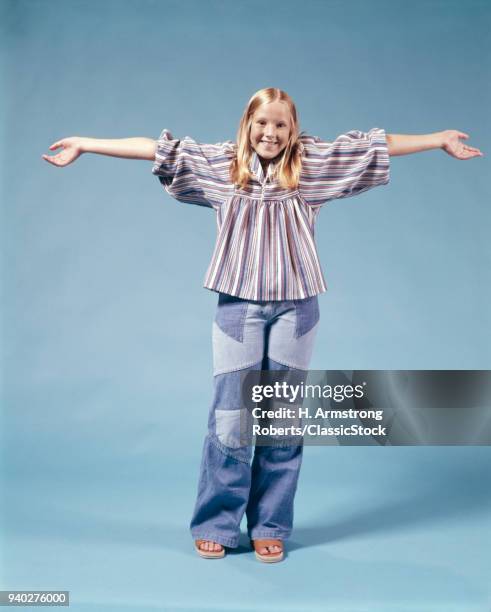 1970s BLOND TEEN GIRL STANDING FULL LENGTH WEARING PATCHWORK BELL BOTTOM DENIM BLUE JEANS ARMS STRETCHED OUT LOOKING AT CAMERA