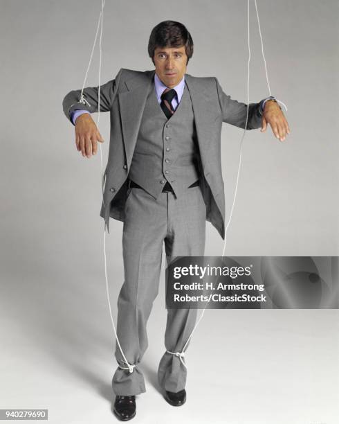 1970s BUSINESSMAN SUSPENDED ON PUPPET STRINGS LOOKING AT CAMERA