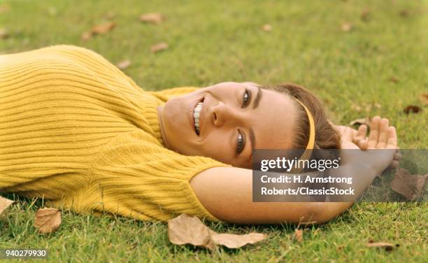 1960s SMILING YOUNG BRUNETTE WOMAN LYING IN GRASS AUTUMN LEAVES LOOKING AT CAMERA WEARING YELLOW POOR BOY SWEATER AND HEADBAND