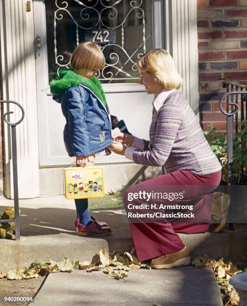 1970s MOTHER ZIPPING DAUGHTERS JACKET AS SHE LEAVES FOR SCHOOL HOLDING LUNCH BOX