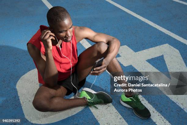 male athlete sitting on all-weather running track and listening to music on smart phone, barcelona, spain - all weather running track fotografías e imágenes de stock